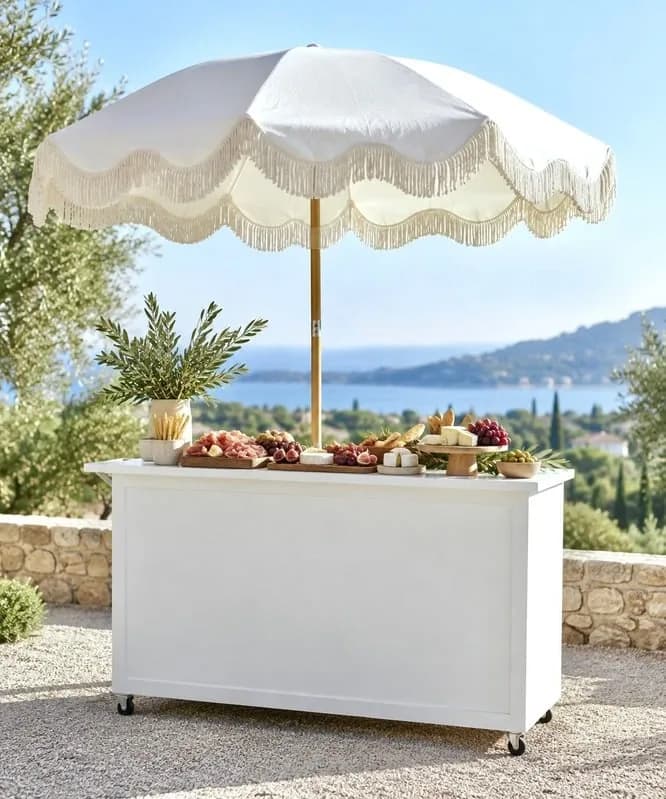 A white cart under a fringed parasol, laid with fruit in a garden
